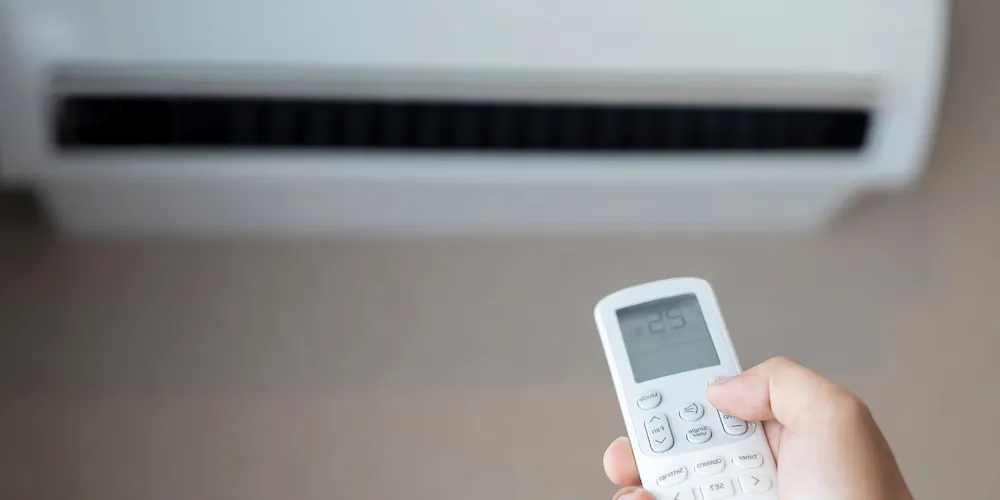 A close-up shot of a person’s hand holding a remote control pointed toward an air conditioning unit. The remote screen shows a temperature setting of 25°C.