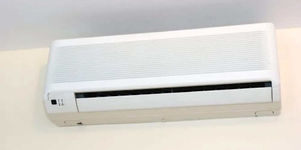 A front view of a compact white air conditioning unit installed high on a wall. The unit has a textured upper grille for air intake and a lower vent for cooling.