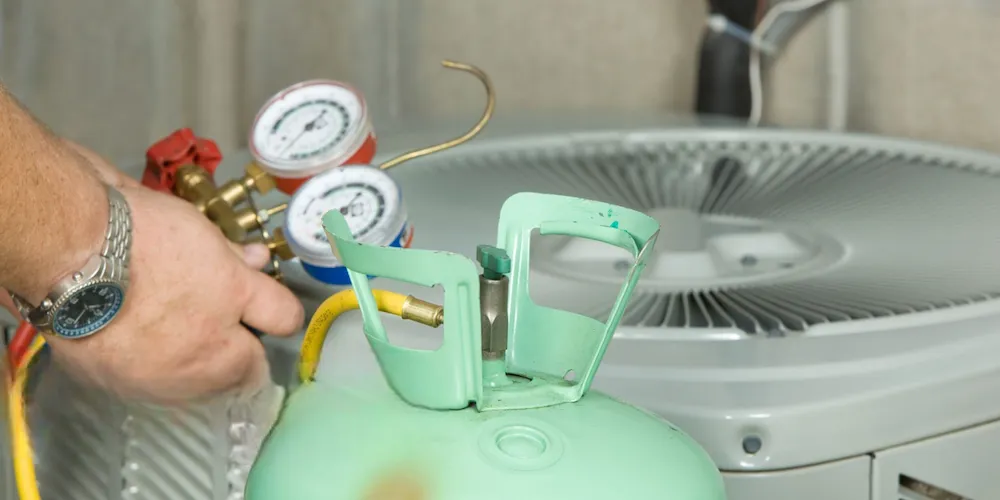 A close-up of an HVAC technician’s hands holding a manifold gauge set (red and blue gauges) connected to a light green refrigerant tank and a grey outdoor AC unit.