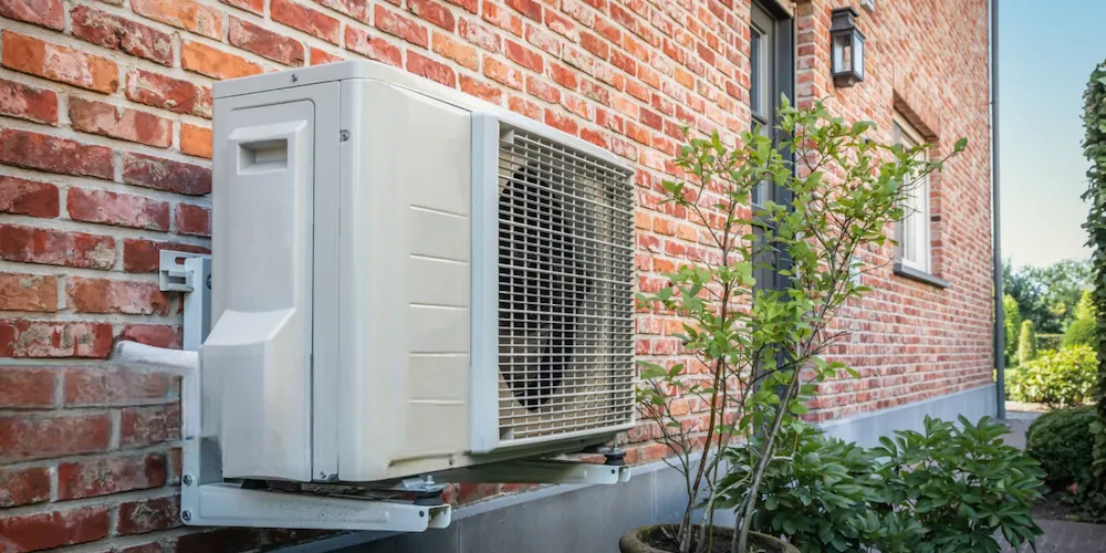 A white, rectangular outdoor air conditioning or heat pump unit mounted on a red brick exterior wall with metal brackets. A small potted plant and green foliage are visible at the base of the wall.