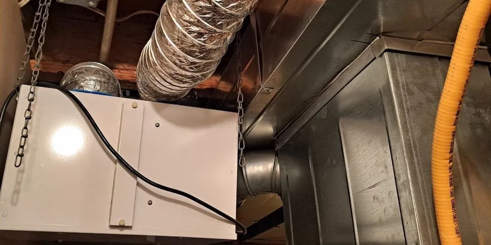 An indoor view of a white rectangular air exchange unit suspended from wooden ceiling joists by metal chains. It is connected to a large silver flexible duct and various metal HVAC trunk lines. A yellow flexible gas line and electrical wiring are also visible.