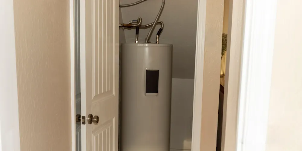 A tall, cylindrical white water heater located inside a small closet or utility room, with flexible silver water lines connected at the top.