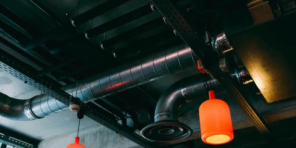 Dimly lit interior with exposed ceiling infrastructure, featuring cylindrical metal ventilation ducts, electrical conduits, and two hanging orange pendant lights.