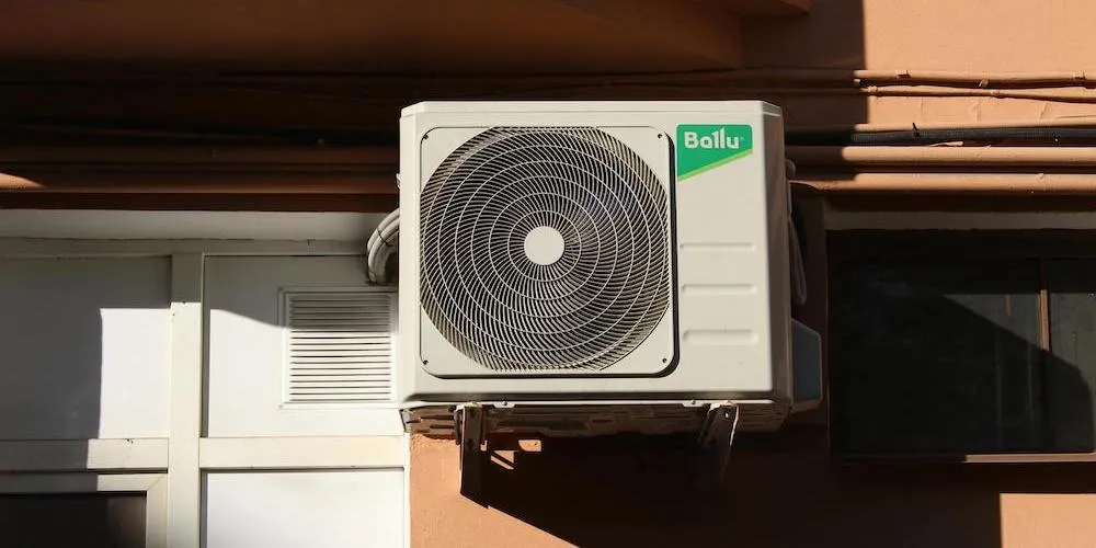 A medium shot of a white Ballu brand outdoor air conditioning compressor mounted to an exterior tan wall with metal brackets. The unit features a large circular fan grille and is positioned next to a window and a white door with a small square vent.